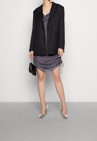 Sort oversized blazer, grå draperet kjole med sidesløjfer, sort håndtaske og sølvfarvede høje hæle. Glatte teksturer og skræddersyet form.