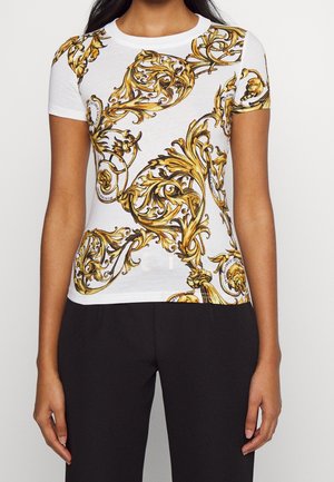 Femme portant une chemise blanche à manches courtes avec un imprimé baroque doré complexe associée à un pantalon noir uni.