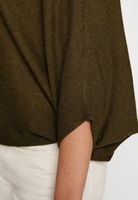 Olijfgroen, geribbelde gebreide top met brede, gedrapeerde mouwen en een ronde zoom. Zachte textuur en casual pasvorm. Witte broekmanchetten zijn zichtbaar.