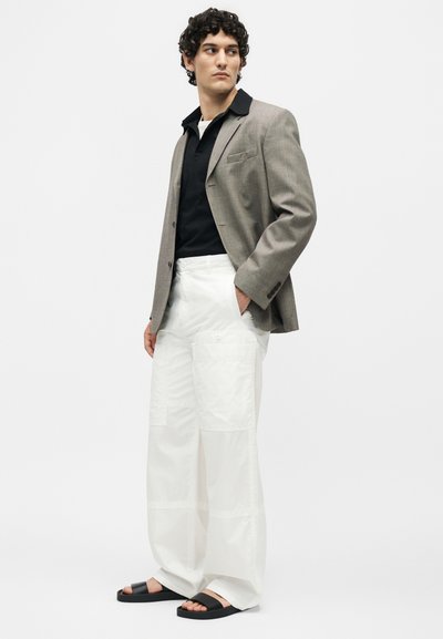 Άνδρας με σγουρά μαλλιά που φοράει γκρί σακάκι, μαύρο πουκάμισο, λευκό παντελόνι φαρδιάς γραμμής και μαύρα σανδάλια, που στέκεται μπροστά σε λευκό μονόχρωμο φόντο.