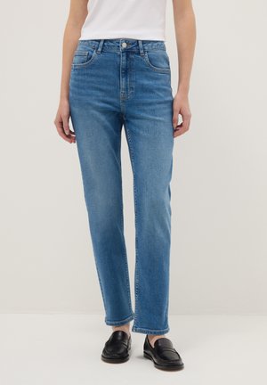 Person trägt mittelhohe, gerade geschnittene blaue Jeans mit ausgewaschenem Effekt, weißes Oberteil und schwarze Loafers, steht vor einem schlichten Hintergrund.