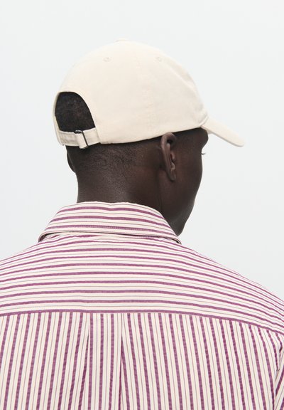 Šviesiai rudos spalvos beisbolo kepurė, pagaminta iš audinio, su užtrauktuku gale. Asmuo vilki dryžuotą marškinėlį šviesios grietinėlės ir purpurinės spalvų derinyje su apykakle.