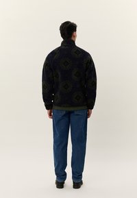 Ζακέτα φλις σε ναυτικό μπλε και πράσινο με σχέδιο, με ψηλό γιακά, συνδυασμένη με μπλε τζιν παντελόνι. Διαθέτει άνετη εφαρμογή και υφή υφάσματος με ανάγλυφα στοιχεία.