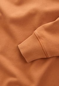 Nærbillede af en manchet på en langærmet beklædningsgenstand i brændt orange med ribstrikket tekstur og glat stofkrop.