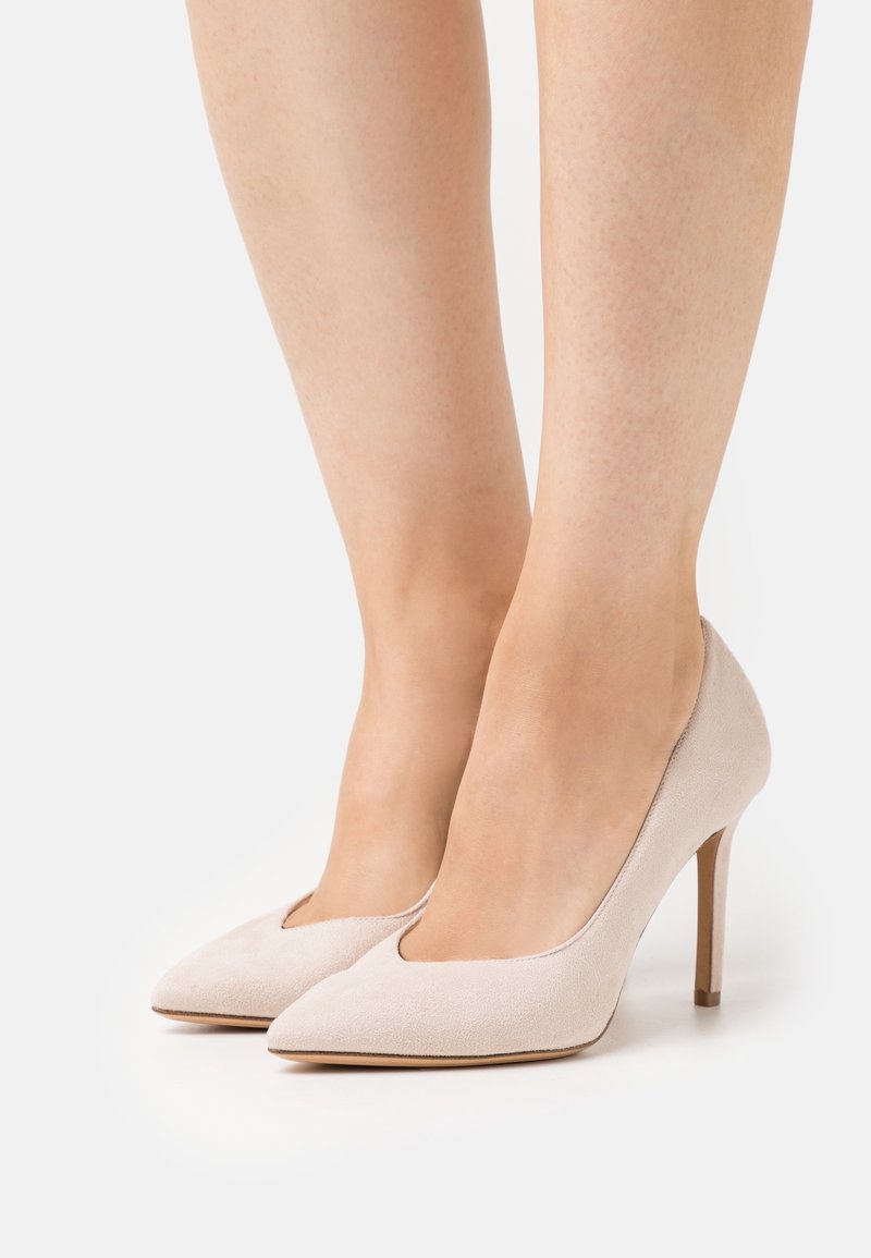 Jambes portant des escarpins à bout pointu en daim beige avec des talons aiguilles fins sur fond blanc.