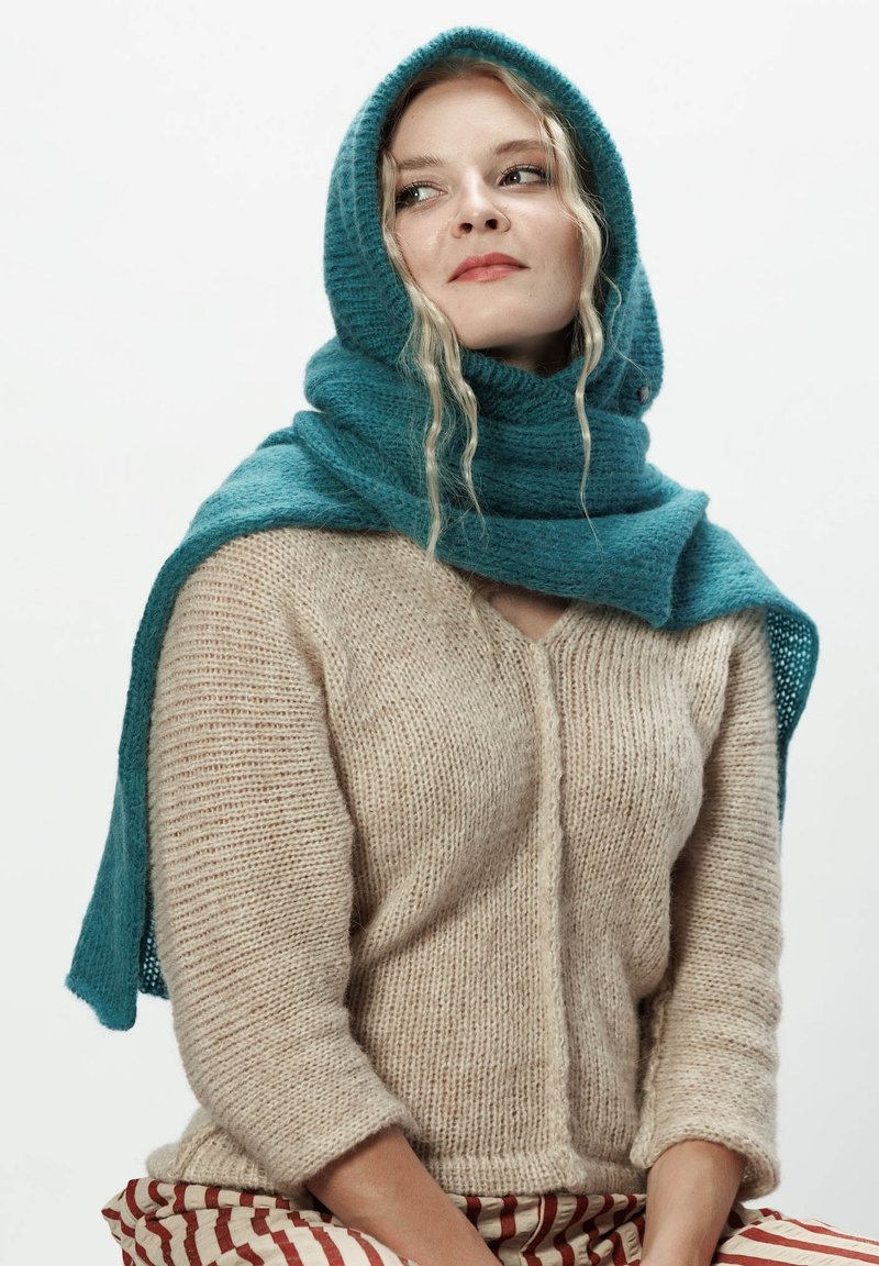 Kobieta w beżowym swetrze dzianinowym, turkusowym kapturze i spodniach w czerwono-białe paski, patrząca lekko w lewo na jednolitym tle.