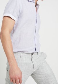 Camicetta di lino lilla chiaro a maniche corte con bottoni frontali e risvolti, abbinata a pantaloni grigi chiari con texture e passanti per cintura.