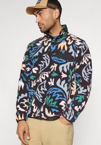Chaqueta tipo polar con estampado de hojas multicolores sobre un fondo oscuro, con un bolsillo en el pecho negro y cuello negro. Textura ligera y suave.