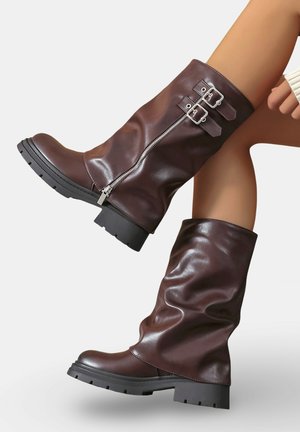 Bottes en cuir marron jusqu'au genou avec une finition lisse, fermeture éclair sur le côté et deux accents de boucle. Elles présentent une semelle en caoutchouc épaisse avec un motif de traction.