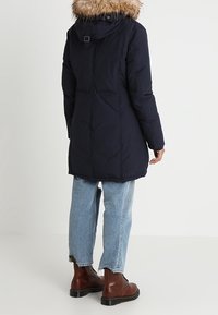 Marinblå lång parka med avtagbar pälsmössa, kviltad design och sidofickor, parat med ljusblå jeans och bruna snörstövlar.
