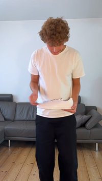 Sneaker bianco tenuto in mano, tomaia in pelle liscia, punta rotonda, suola piatta, occhielli argentati. Pantaloni neri e divano grigio visibili sullo sfondo.