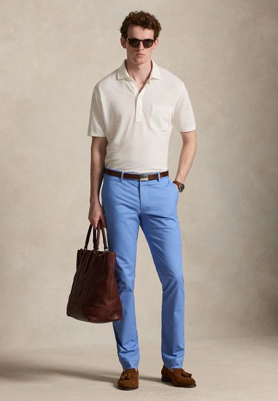 Άνδρας που φορά λευκό πόλο με κοντά μανίκια, μπλε παντελόνια, καφέ ζώνη και παπούτσια, κρατώντας μεγάλη καφέ δερμάτινη τσάντα, στέκεται μπροστά σε μπεζ φόντο.