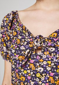 Bluzka w kwiatowy wzór z bufiastymi rękawami. Posiada detal w postaci węzła przy dekolcie, wykonana z miękkiego materiału w odcieniach fioletu, pomarańczu i żółci.