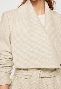 Abrigo cruzado beige en tejido texturizado acanalado. Con cuello ancho y cintura con cinturón, mangas largas y una apariencia suave y acogedora.