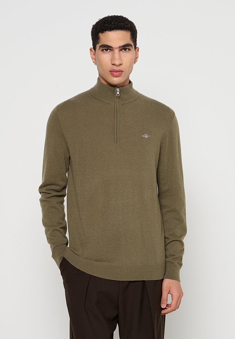 Oliwkowa rozpinana sweterka z miękkiego materiału, z wysokim kołnierzem, długimi rękawami i subtelnym emblematem na lewej piersi.