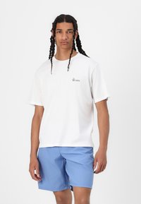 Camiseta de algodón blanca con mangas cortas, que presenta un pequeño logo negro. Combinada con pantalones cortos azules, ambos sobre un fondo blanco.