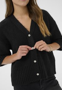 Black knitted cardigan with short sleeves, featuring a V-neck design and decorative silver flower buttons. Ribbed hem for texture contrast.