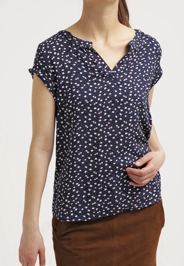 Femme portant un chemisier bleu marine avec de petits motifs floraux blancs et roses et une jupe marron, main posée sur la hanche.