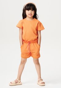 Combinaison en tricot orange avec manches courtes, taille élastique et poches. Fini avec un cordon rose et une bordure en coquilles. Porté avec des sandales beiges.
