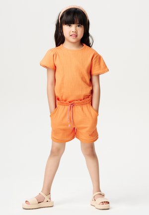 Combinaison en tricot orange avec manches courtes, taille élastique et poches. Fini avec un cordon rose et une bordure en coquilles. Porté avec des sandales beiges.