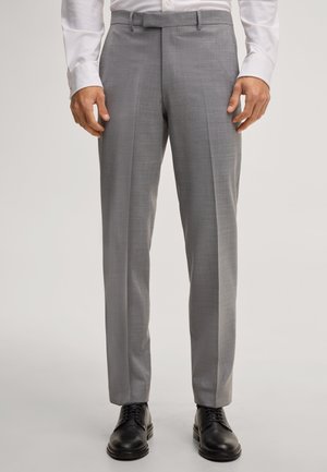 Man die lichtgrijze op maat gemaakte pantalon draagt, wit overhemd en zwarte nette schoenen, staand met ontspannen handen langs zijn zij.