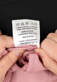 Etichetta nera attaccata a un tessuto rosa. L'etichetta mostra le istruzioni per la cura, i dettagli del prodotto e le informazioni del produttore. Il testo è in più lingue.