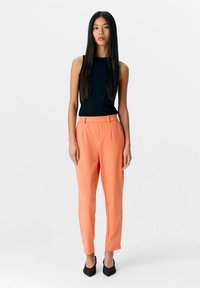 Haut sans manches noir associé à des pantalons orange ajustés et des escarpins noirs à bout pointu. Texture de tissu lisse, design ajusté et coupe droite.