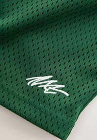 Grön atletisk nätväv med diamantformade perforeringar, med en vit broderad logotyp. Slät textur med en andningsbar design.