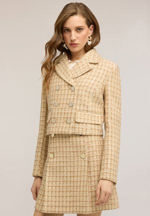 Femme portant un blazer beige à carreaux croisé et une jupe trapèze assortie avec des boutons dorés et des boucles d'oreilles en forme de cœur.