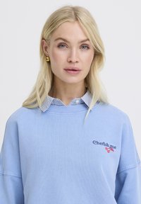 Blonde vrouw met blauwe ogen draagt gouden bolvormige oorbellen, een blauwe trui met "Cherish me" en een rode kersenborduursel, over een gestreept overhemd met kraag.