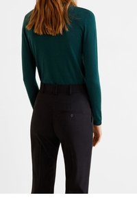 Femme portant un haut à manches longues vert foncé rentré dans un pantalon noir ajusté taille haute, se tenant de dos face au spectateur.
