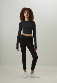 Top cropped preto de mangas longas e jeans skinny pretos, combinados com sapatilhas brancas. Modelo posicionado em um fundo neutro.