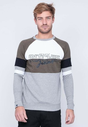 Jeune homme aux cheveux châtain clair portant un sweat-shirt bicolore avec le texte "Ritchie Jeans", debout devant un fond gris clair uni.