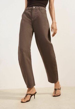 Osoba nosiaca hnedé nohavice s vysokým pásom a voľným strihom a čierne sandále na podpätku, stojaca na jednoduchom svetlom pozadí.