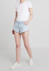 Persona vestida con una camiseta blanca simple, pantalones cortos de mezclilla azul claro arremangados y zapatillas blancas, de pie con una mano en el bolsillo.