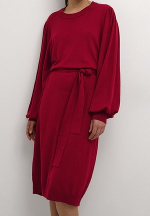 Femme portant une robe rouge longueur genou avec de longues manches bouffantes et une ceinture en tissu nouée à la taille, debout contre un mur blanc.