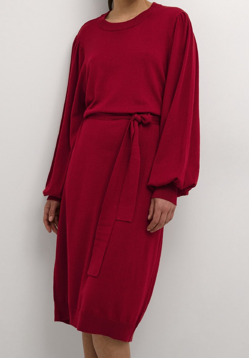 Femme portant une robe rouge longueur genou avec de longues manches bouffantes et une ceinture en tissu nouée à la taille, debout contre un mur blanc.