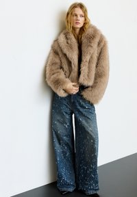 Jonge vrouw die een oversized tan nep bontjas en wijd uitlopende blauwe spijkerbroek draagt, staand tegen een witte muur.