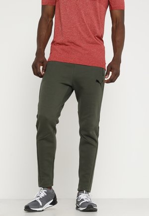 Homme portant un jogging Puma vert olive, un t-shirt rouge chiné et des chaussures de sport grises, debout sur un fond blanc uni.