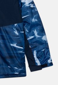 Sezione di giacca blu navy con un tessuto texture, caratterizzata da un motivo ondulato in toni di blu più chiari. Include un dettaglio di polsino in Velcro.