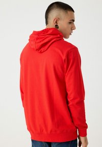 Sudadera roja de tejido suave, con capucha, mangas largas y puños acanalados. La vista trasera muestra una textura lisa sin patrones.