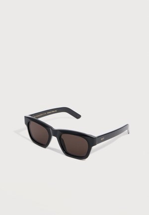 Lunettes de soleil rectangulaires noires avec des verres foncés et des branches fines, placées en angle sur une surface blanche.