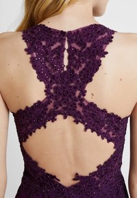 Fioletowa haftowana sukienka z kwiatowym koronkowym wzorem, z wycięciem na plecach i błyszczącymi akcentami, podkreślająca misterną szczegółowość i teksturę.