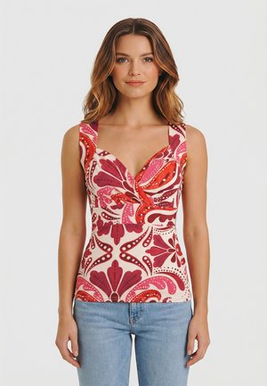 Donna che indossa una maglietta senza maniche, aderente, con motivo floreale rosso e rosa e jeans azzurri, in piedi davanti a uno sfondo bianco semplice.