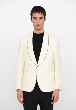 Cremfarbener Blazer mit schwarzem Reverspiping, taillierte Passform, einreihiger Knopfverschluss und Schalkragen. Das Modell trägt einen schwarzen gerippten Pullover darunter.