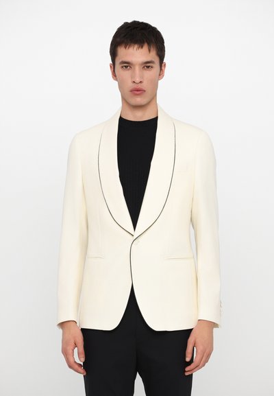 Blazer crème avec galon noir sur le revers, design ajusté, fermeture à un bouton et revers crantés. Le modèle porte un pull côtelé noir en dessous.