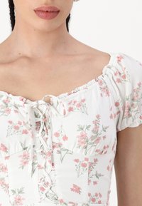 Nainen, jolla on päällään valkoinen, lyhythihainen paita, jossa on vaaleanpunaisia kukkakuoseja ja nauhakuja. Kuvassa näkyy hänet kaulasta vyötäröön.