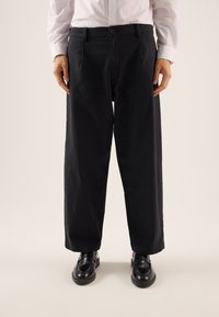 Pantaloni in cotone neri con una vestibilità rilassata, gambe dritte e tasche laterali. Indossati con scarpe nere lucide, in abbinamento a una camicia bianca.
