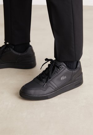 Zapatillas negras de cuero Lacoste con cordones negros, usadas con pantalones y calcetines negros sobre un suelo de color claro.