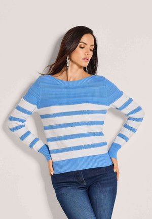 Vrouw met lang donker haar, gekleed in een blauw-wit gestreepte trui en donkere spijkerbroek, poseert met handen in de zij tegen een lichte muur.